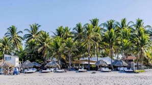 Egy pálmafákkal, napozóágyakkal és szalmatetős építményekkel ellátott strand.