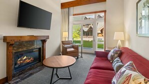 A living room with a fireplace, a television, a sofa, a chair, and a view of a building through a glass door. - Spruce Peak Lodge | One Bedroom | Alpine Getaway (Stowe)
