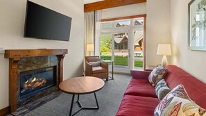 Un salon avec une cheminée, un téléviseur, un canapé, un fauteuil et une vue sur un immeuble à travers une porte vitrée.