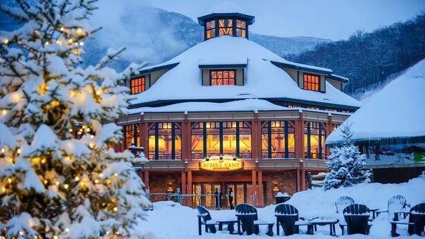 En stuga med ett snötäckt tak, flera fönster och en skylt med texten ”Spruce Camp East Lodge”.