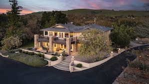A two-story house with a balcony, surrounded by landscaped gardens and a hilly landscape in the background. - El Pomar Estate - Main House by Avantstay Pool + Hot Tub + Firepit + Outdoor Dining + Vineyard Views (Templeton)