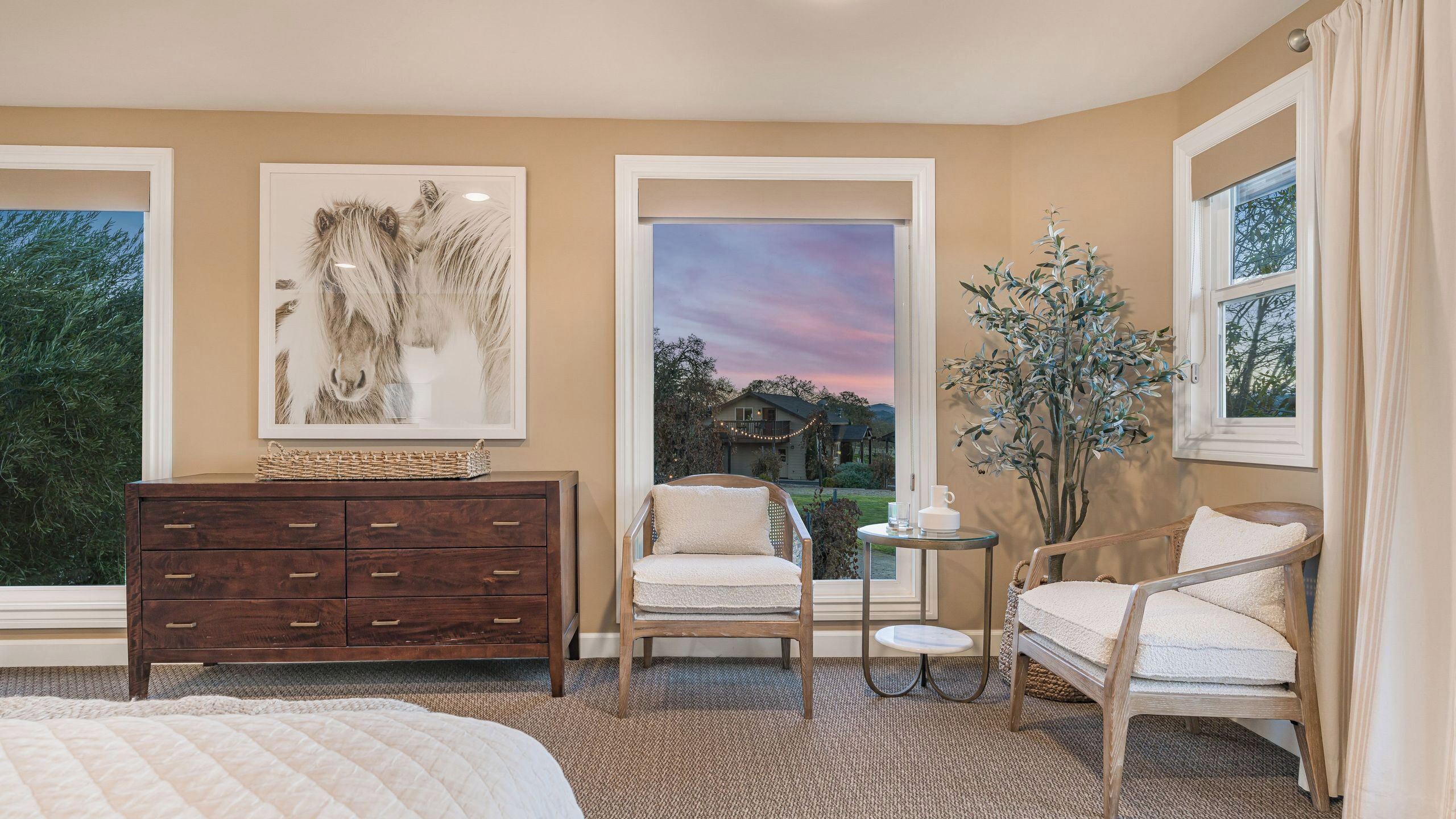A bedroom with a large bed, a wooden dresser, two chairs, a small table, and a view of a house through a window.