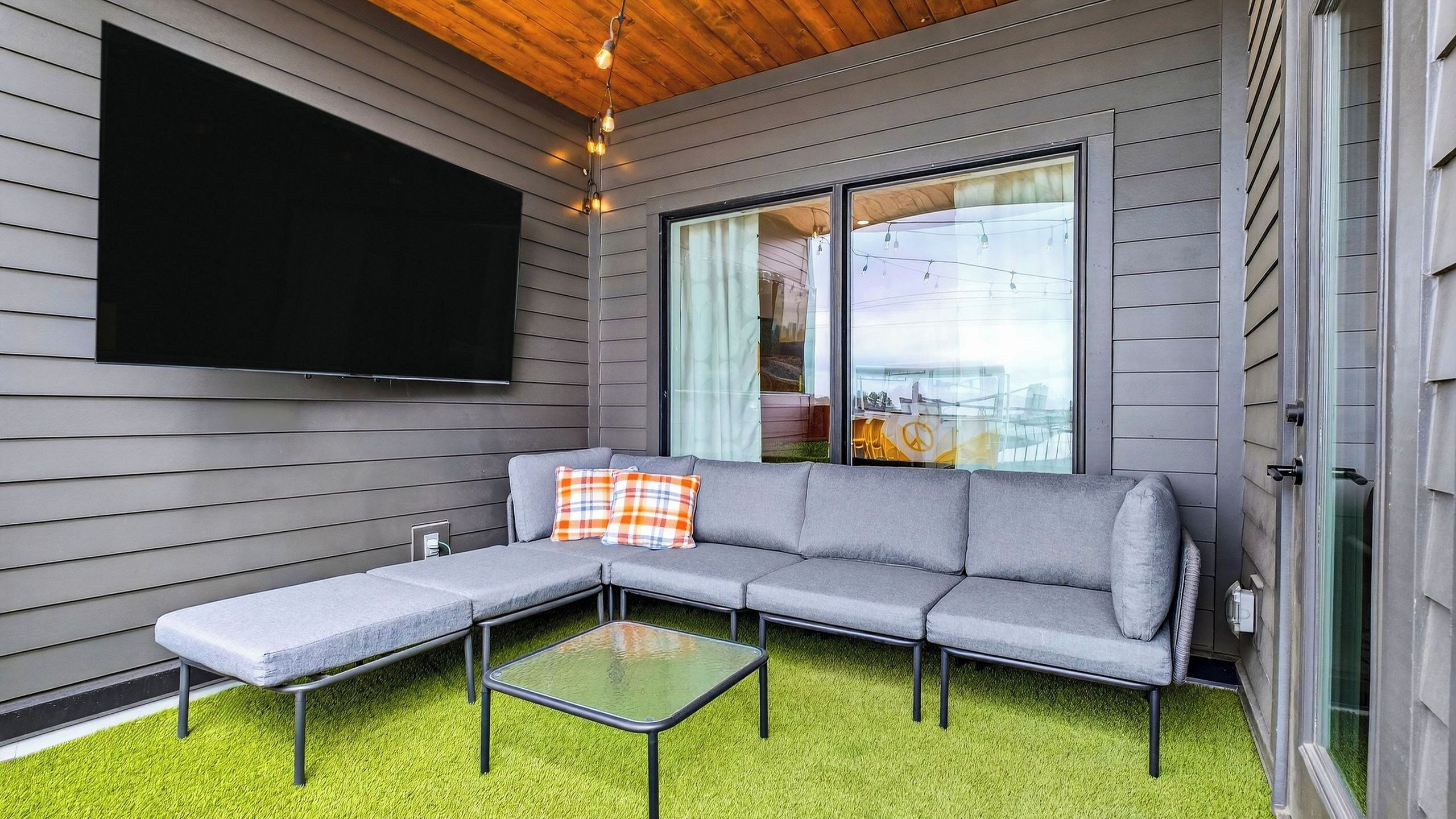 A modern outdoor living space with a grey sectional sofa, a glass coffee table, and a flat-screen TV mounted on the wall.