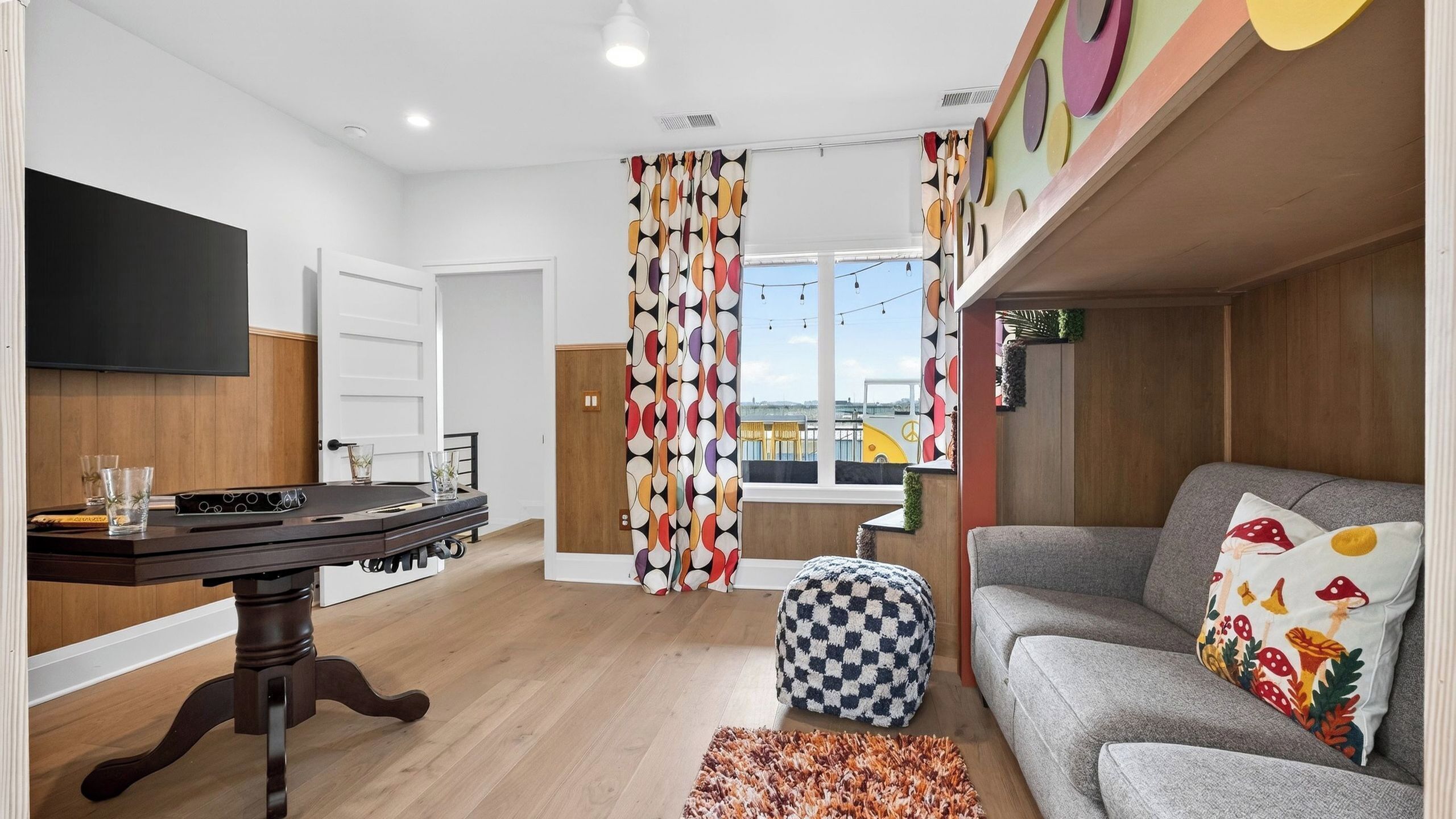 A living room with a sofa, a table with glasses, a checkered ottoman, and a TV mounted on the wall.