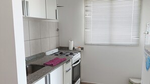 Une cuisine avec des meubles blancs, une cuisinière à gaz, un four encastré et une fenêtre avec des stores.