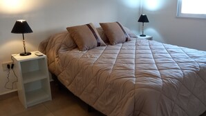 Un lit bien fait, recouvert d’une couette beige, avec deux lampes de chevet, une table de nuit et une fenêtre laissant entrer la lumière du jour.