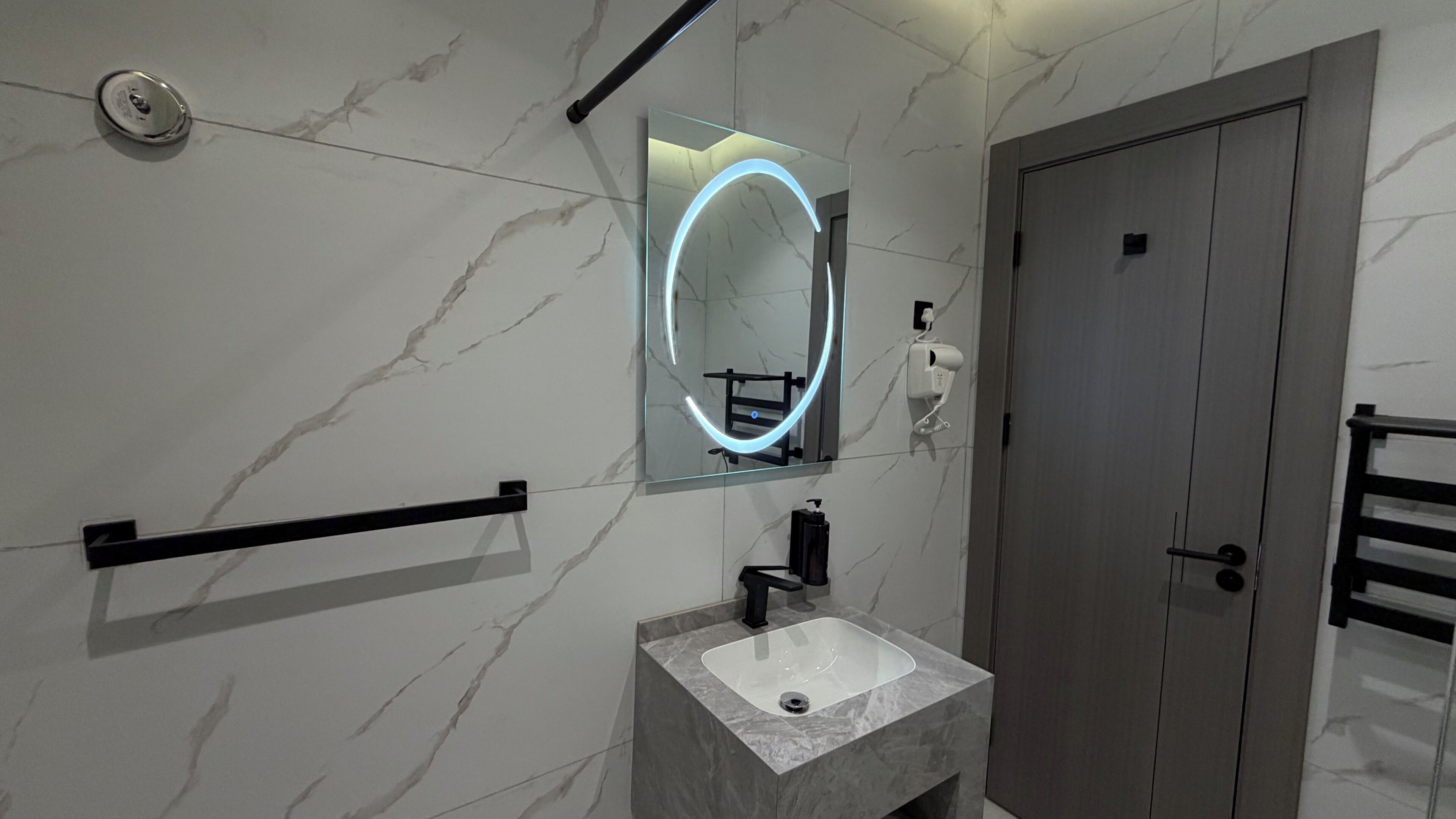 Un baño moderno con pared de mármol, un espejo grande con iluminación envolvente, un toallero negro, un lavabo con grifo negro y una puerta.