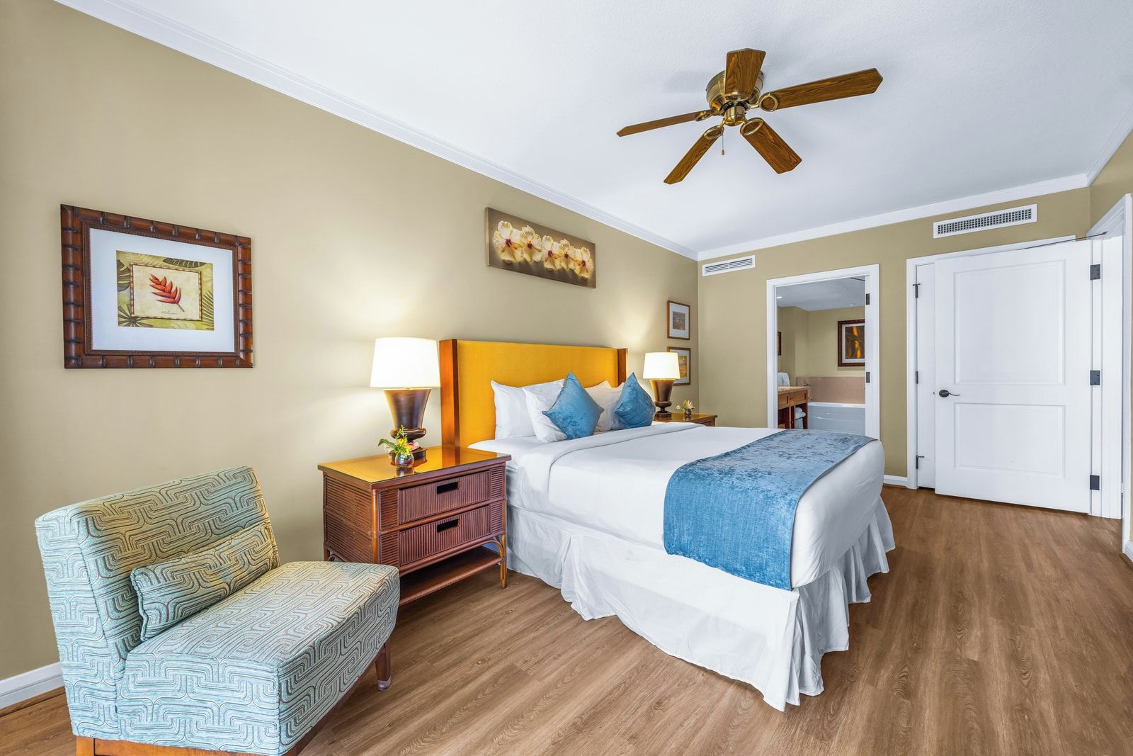 Une chambre d’hôtel avec un lit, une chaise, une table de chevet, un ventilateur de plafond et une porte.