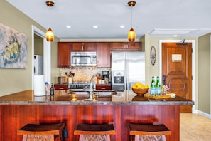 A kitchen with wooden cabinets, stainless steel appliances, a central island with bar stools, and pendant lighting. - Honua Kai | Ocean View 7 BR, Sleeps 20 | Car Incl. w/ 6+ Nights | HKK ML-3356 by KBM (Lahaina)