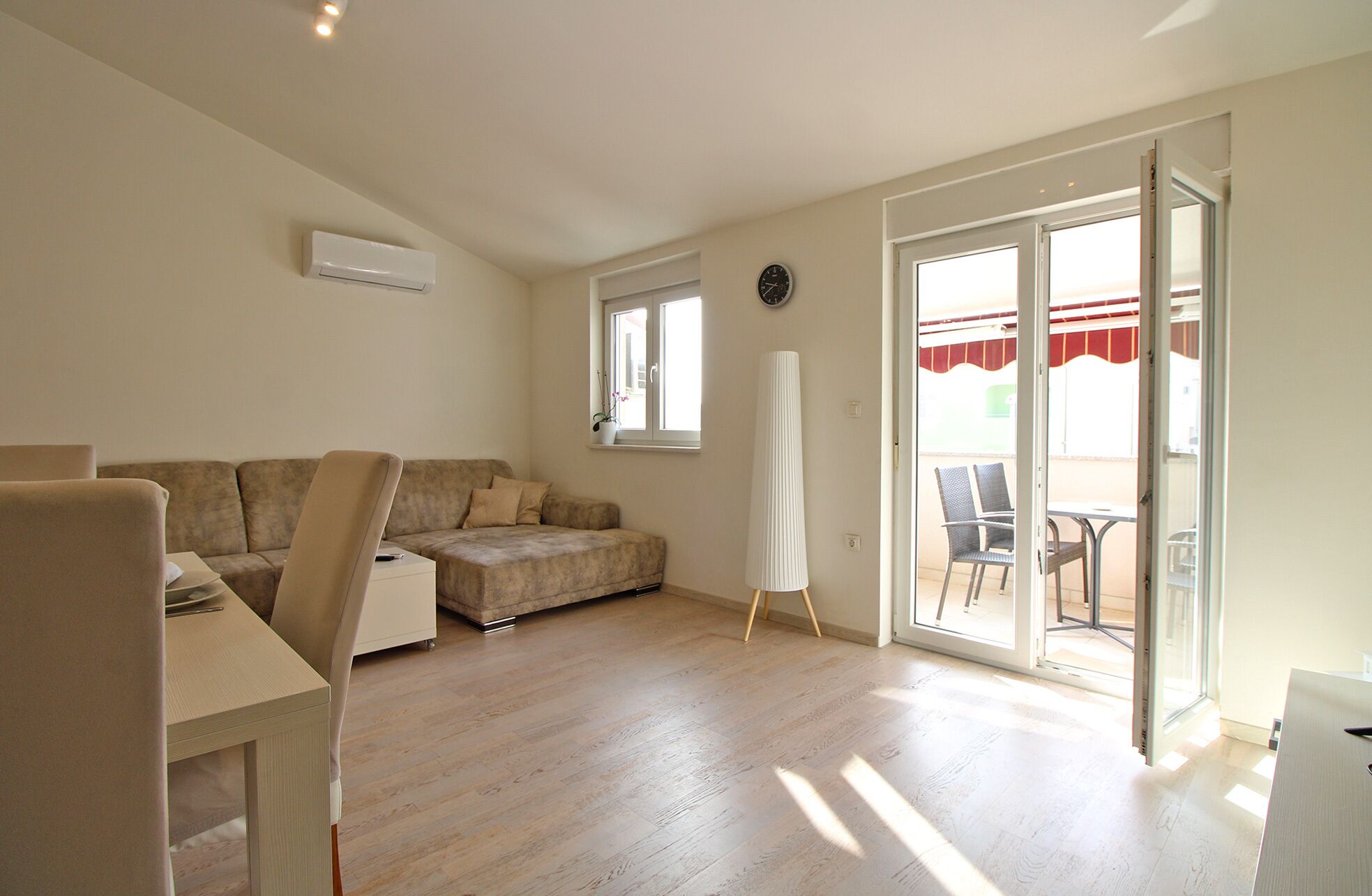 A spacious living area with a sofa, a small table, a standing lamp, and a sliding glass door leading to an outdoor seating area.