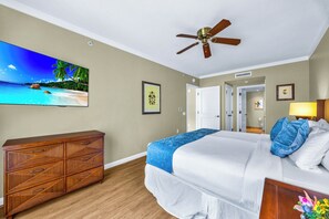 Ein Hotelzimmer mit einem Bett, einem Holzkoffer, einem Deckenventilator, einem gerahmten Bild und einer Tür zu einem weiteren Raum.