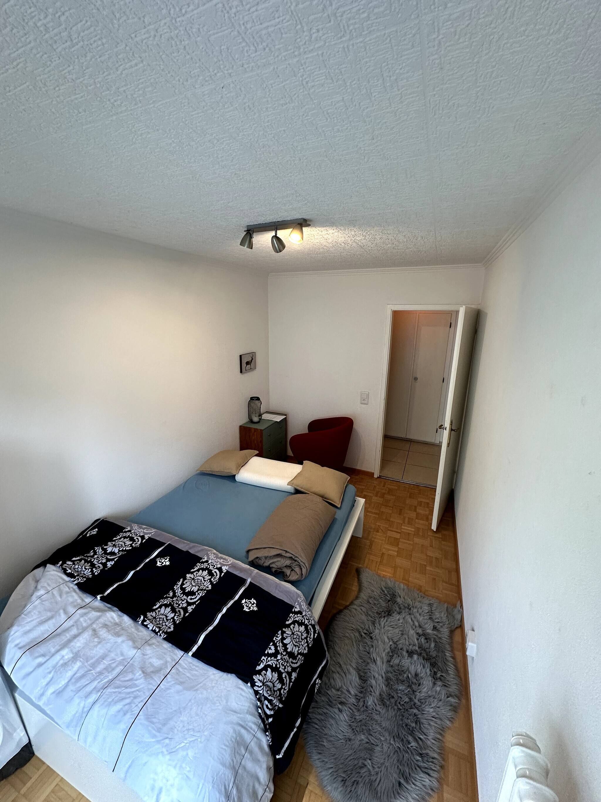 A bedroom with a bed, a red chair, a small table, a picture on the wall, and a door leading to another room.