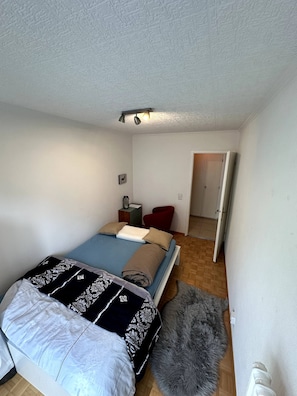 Une chambre à coucher avec un lit, une chaise rouge, une petite table, un tableau accroché au mur et une porte donnant sur une autre pièce.