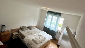 A bedroom with a bed, a nightstand, a window with curtains, and a radiator. - Furnished Apartment in Baden (Baden)
