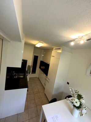 A compact kitchen with white cabinets, a black countertop, and a small sink. There is a white table with a vase of flowers and a piece of paper on it. - Furnished Apartment in Baden (Baden)