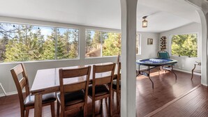 A dining area with a table and chairs, a round table with a board game, and large windows with a forest view. - Honey Bear Cabin (Big Bear Lake)