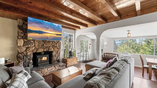 A modern living room with a stone fireplace, a large flat-screen TV, a cozy sofa, and a dining area with natural light. - Honey Bear Cabin (Big Bear Lake)