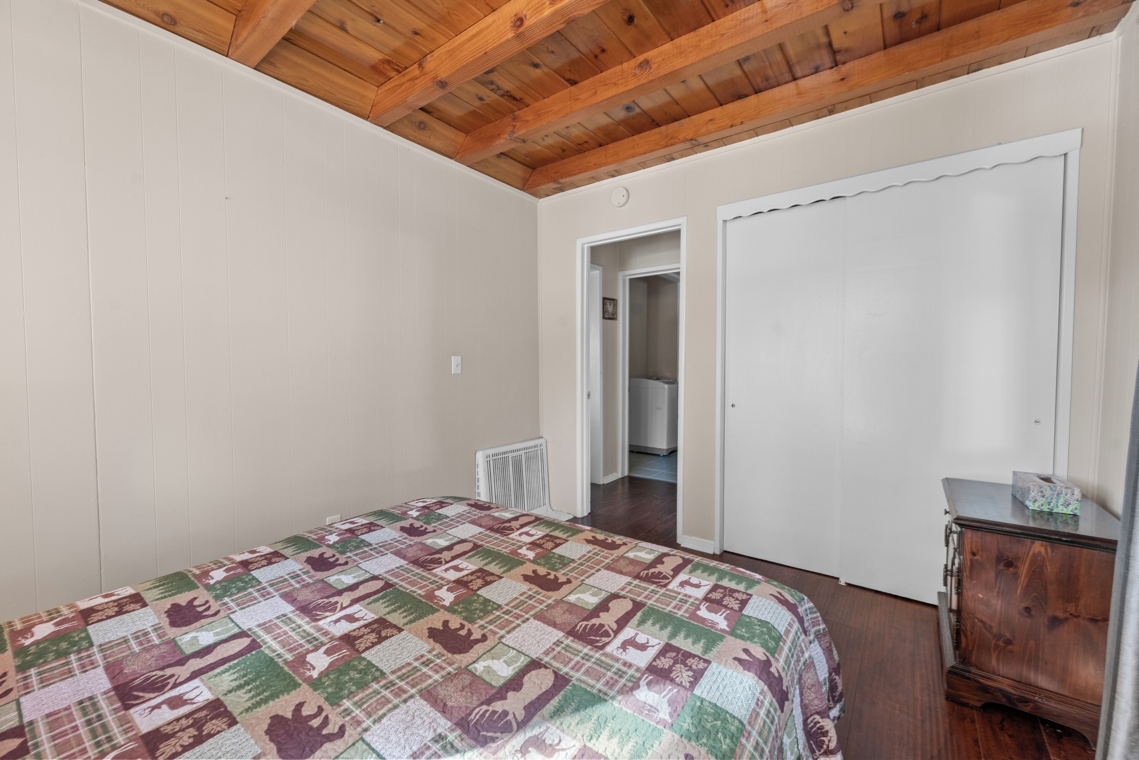 A bedroom with a wooden ceiling, a quilted bedspread, a radiator, a wardrobe, and a door leading to another room.