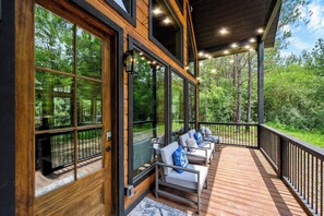 A wooden deck with outdoor seating, large windows, and a view of the forest. - Modern Glass Cabin w/ Hot Tub & Outdoor Lounge (Broken Bow)