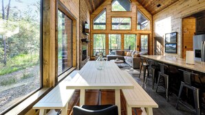 A wooden cabin interior with a dining table, bar area, and large windows offering a forest view. - Modern Glass Cabin w/ Hot Tub & Outdoor Lounge (Broken Bow)