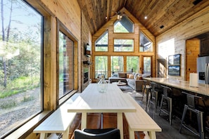A wooden cabin interior with a dining table, bar area, and large windows offering a forest view. - Modern Glass Cabin w/ Hot Tub & Outdoor Lounge (Broken Bow)