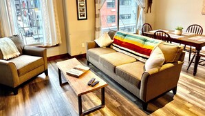 A living room with a sofa, a coffee table, and a dining table with chairs. There is a window with curtains, a framed picture on the wall, and a view of buildings outside.