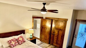 A bedroom with a bed, a nightstand, a lamp, a ceiling fan, wooden wardrobe, and a door.