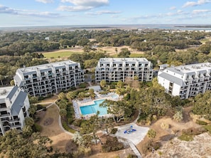 Un hotel de varios pisos con piscina, rodeado de vegetación y un campo de golf al fondo.