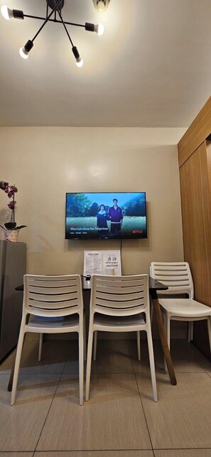 Ein Zimmer mit an der Wand montiertem Fernseher, einer Deckenleuchte und drei weißen Kunststoffstühlen.