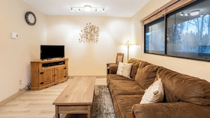 A living room with a wooden TV stand, a brown sofa, a coffee table, and a wall-mounted light fixture. - Marina Cay Condo | Sleeps 4 | Steps from Flathead Lake, Bigfork Village & Glacier Country (Bigfork)
