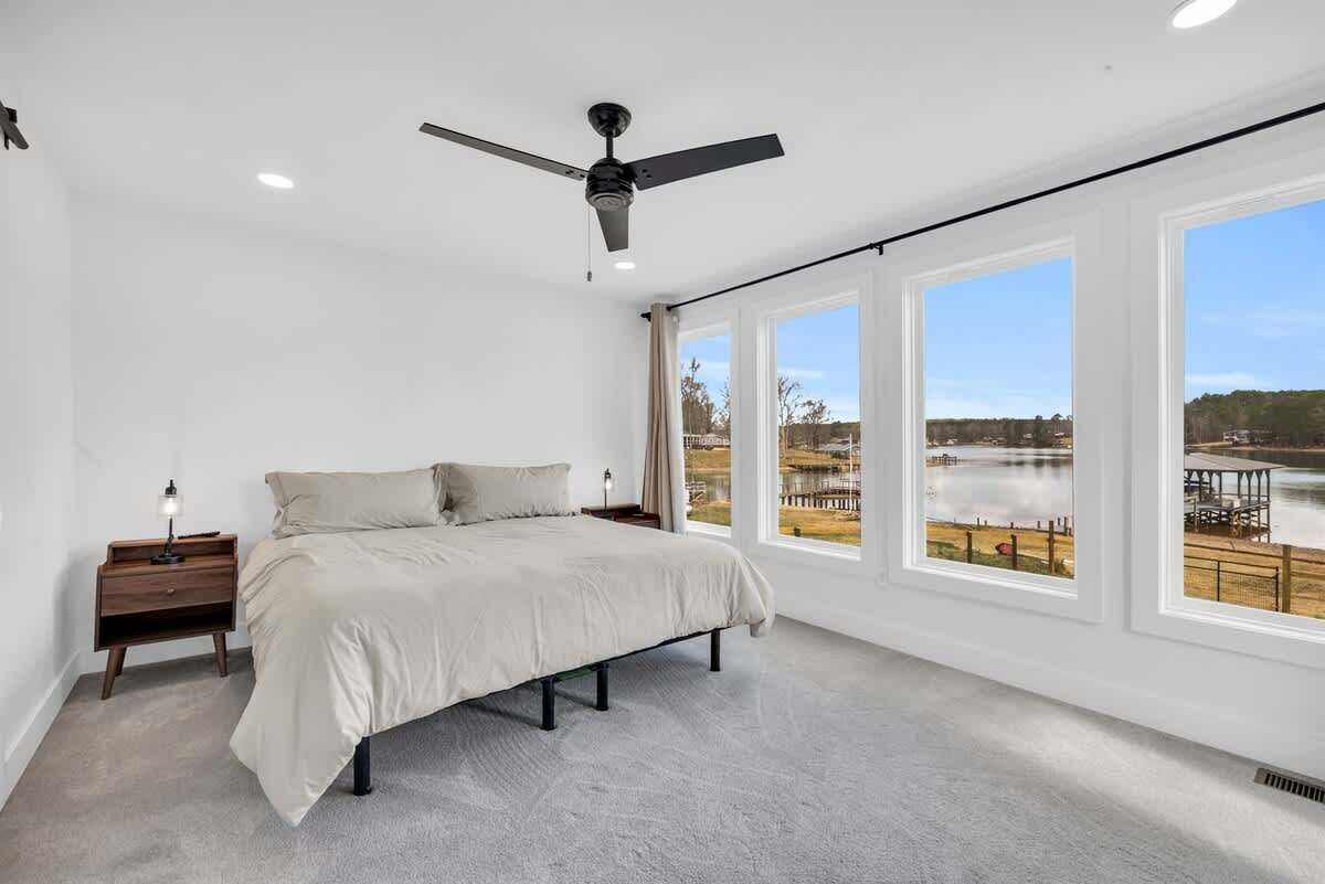 Une chambre à coucher comprenant un lit, un ventilateur de plafond, une table de chevet et de grandes fenêtres donnant sur un plan d’eau et un quai.