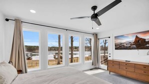 A bedroom with a large bed, a ceiling fan, a wooden dresser, and a view of a lake and trees through large windows.