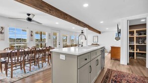 A spacious kitchen with a large island, wooden dining table, and chairs, with a view of the outdoors through large windows.