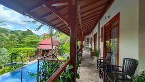 A covered balcony with a pool, outdoor seating, and a view of a red-roofed building amidst lush greenery.