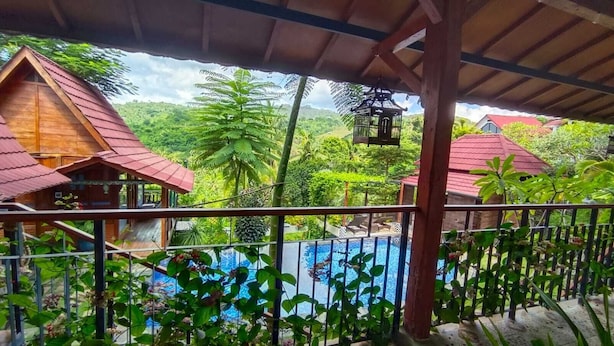 A wooden house with a red roof, a pool, and lush greenery.