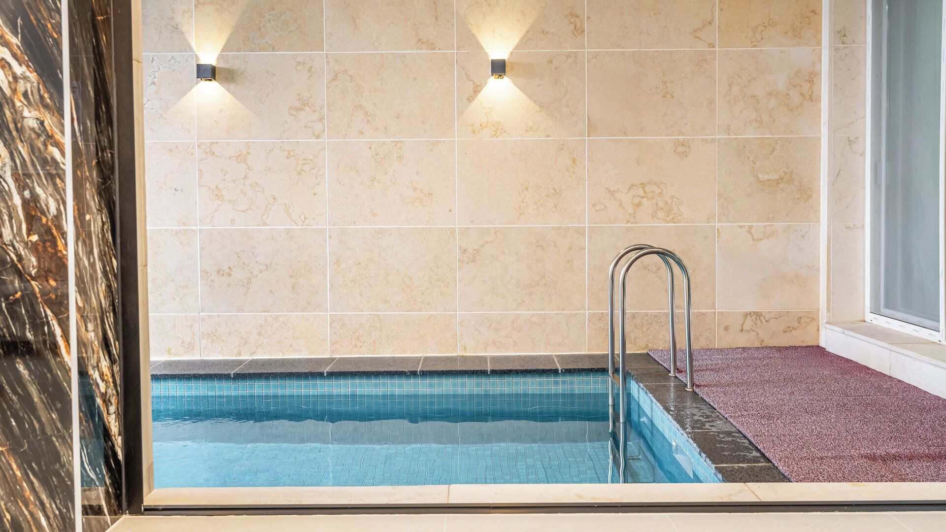 Una piscina con una escalera metálica, paredes de azulejos color beige y dos luces empotradas.