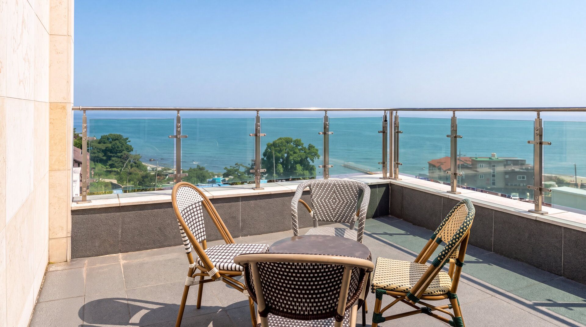 Un balcón con sillas de mimbre y una mesa, con vista a un mar despejado y edificios lejanos.