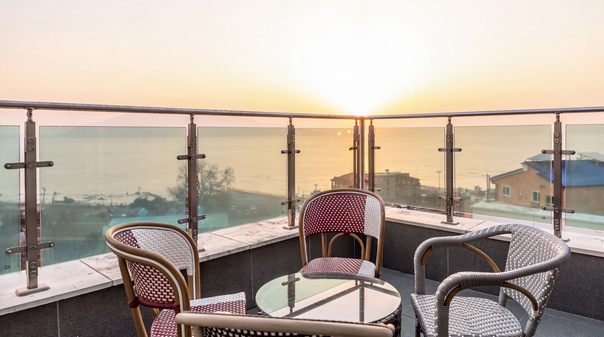 A balcony with wicker chairs and a glass table overlooking a body of water during sunset.
