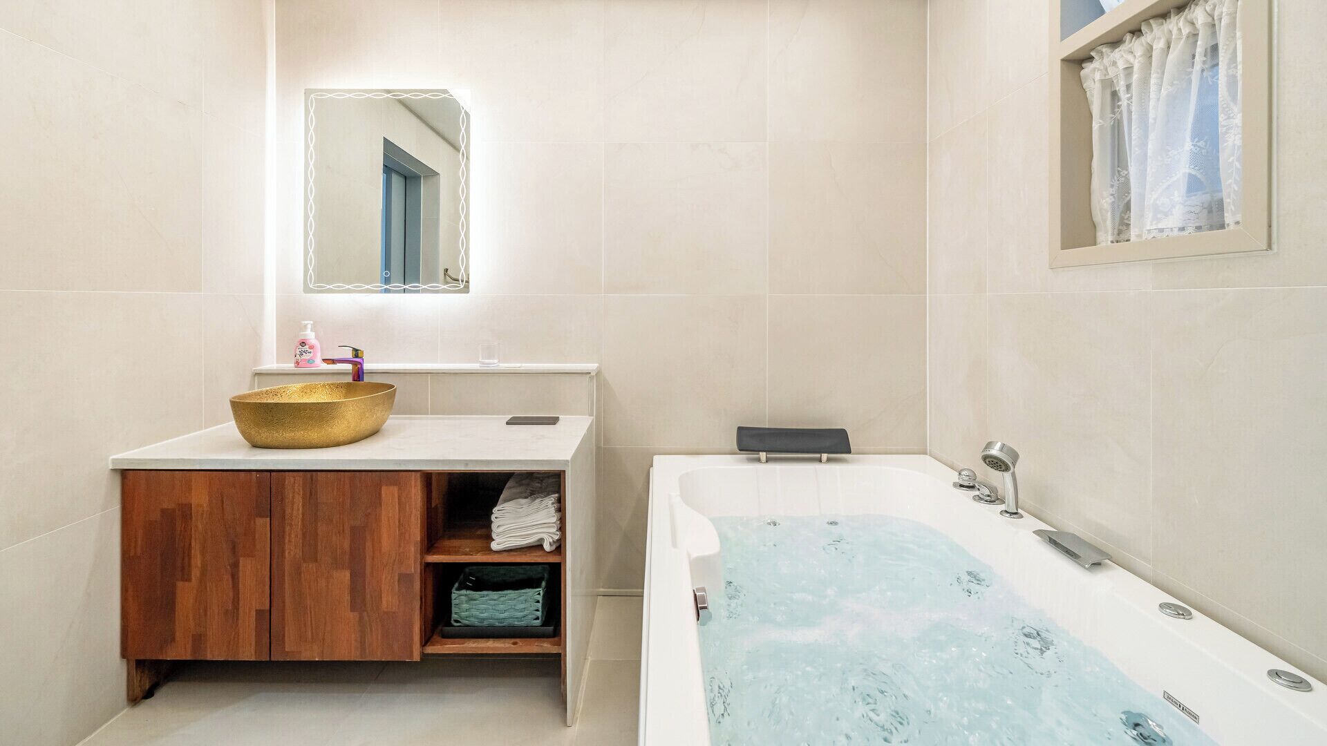 A modern bathroom with a wooden vanity, a gold bowl sink, a large bathtub, and a mirror with a decorative frame.