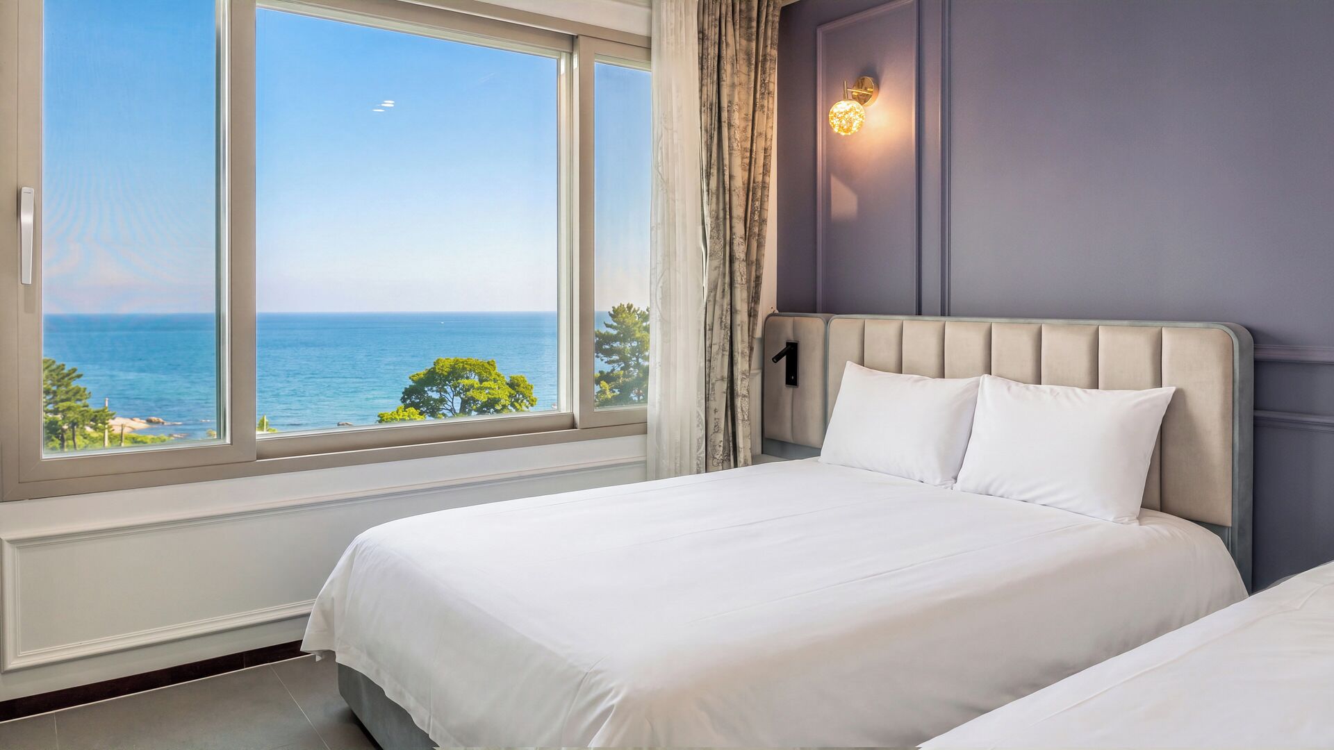 Habitación de hotel con un ventanal que ofrece vistas al mar y a los árboles. Se observa una cama con cabecera y dos almohadas.