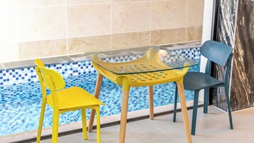 A poolside area with a glass table and two chairs, one yellow and one blue, with a tiled wall in the background.