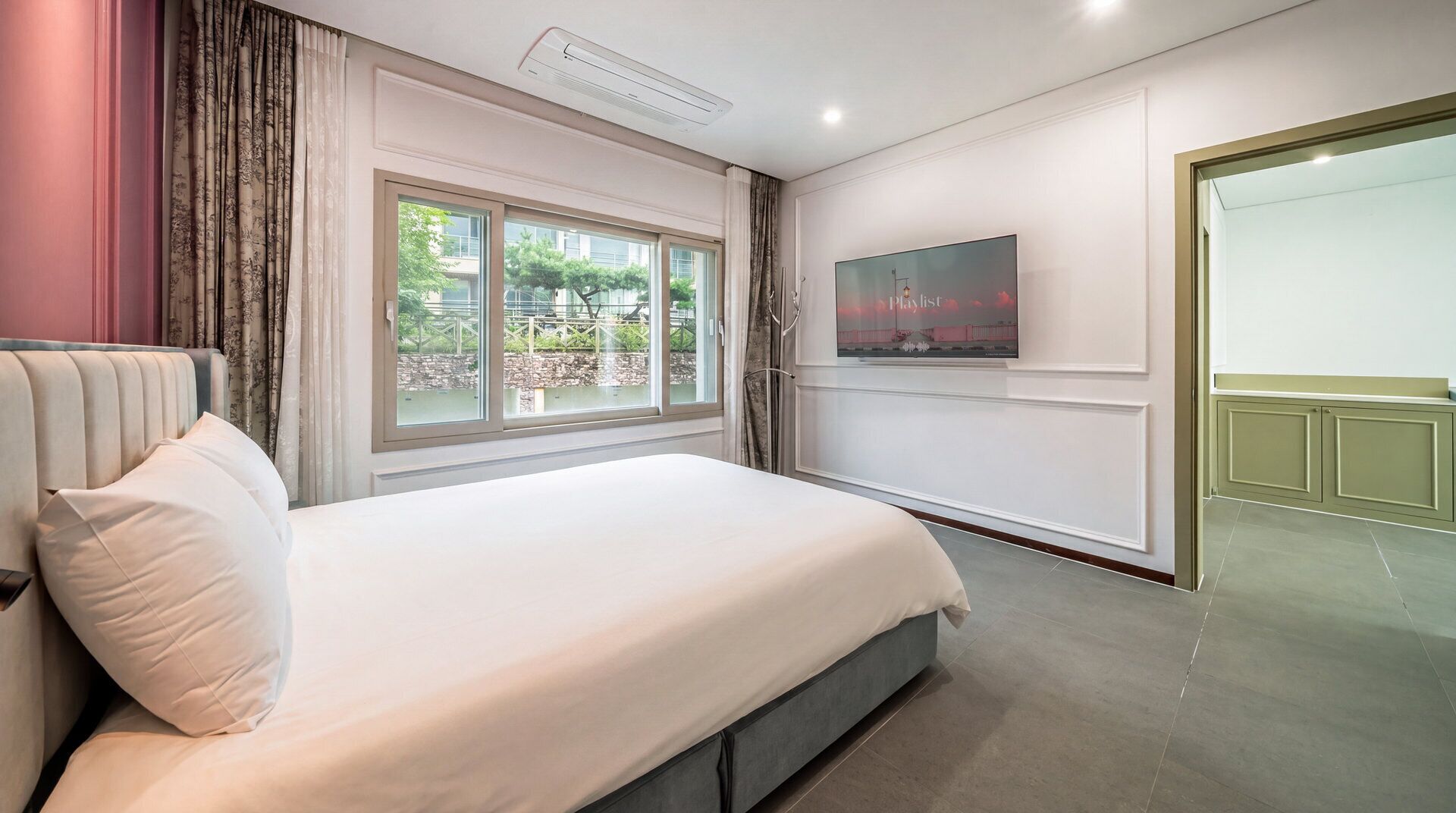 A hotel room with a bed, a TV, a window with curtains, and a door leading to another room.