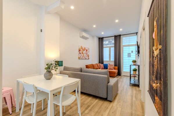 A modern living room with a sofa, a dining table with chairs, a small kitchen area, and a large window with blinds.