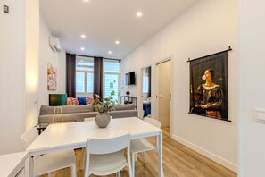A modern living room with a white sofa, a dining table with chairs, a large painting on the wall, and a TV mounted on the wall.