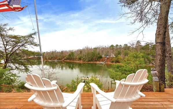 Eine Seeterrasse mit zwei weißen Adirondack-Stühlen, die auf einen ruhigen See blicken, umgeben von Bäumen und einem Steg.