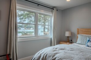 A bedroom with a bed, a nightstand, a lamp, and a window with curtains. - Oyster Haven/Waterfront Retreat on Lanes Creek/Mathews, VA (Hudgins)