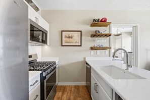 Eine moderne Küche mit einem Edelstahlkühlschrank, einem eingebauten Backofen, einem Gasherd, einer weißen Arbeitsplatte und einem Spülbecken. Über dem Spülbecken befinden sich Holzregale und ein gerahmtes Bild an der Wand.