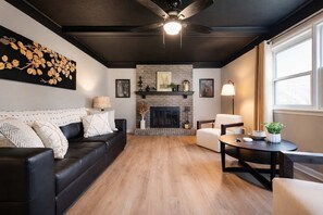 A living room with a fireplace, a black leather sofa, a white armchair, a round table, and a ceiling fan. - Spacious Louisville home with backyard and parking (Louisville)