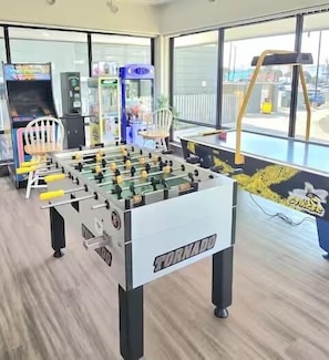 A foosball table labeled 'TORNADO' in a room with large windows, a chair, and a vending machine. - Oceanfront Corner Unit with Pool on Boardwalk (Carolina Beach)