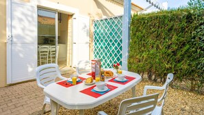 An outdoor dining area with a table set for two, surrounded by white plastic chairs, a white umbrella, and a green lattice fence. - Residence Les Soleillades Et Scarlett (Palavas-les-Flots)